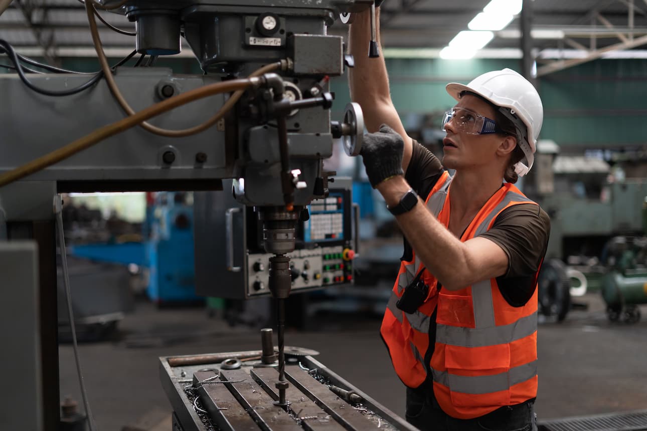 Trabajador dentro de una nave industrial manipulando un torno de perforación con el equipo de protección individual adecuado: casco de protección, gafas de seguridad, chaleco reflectante y guantes de protección anti corte. 